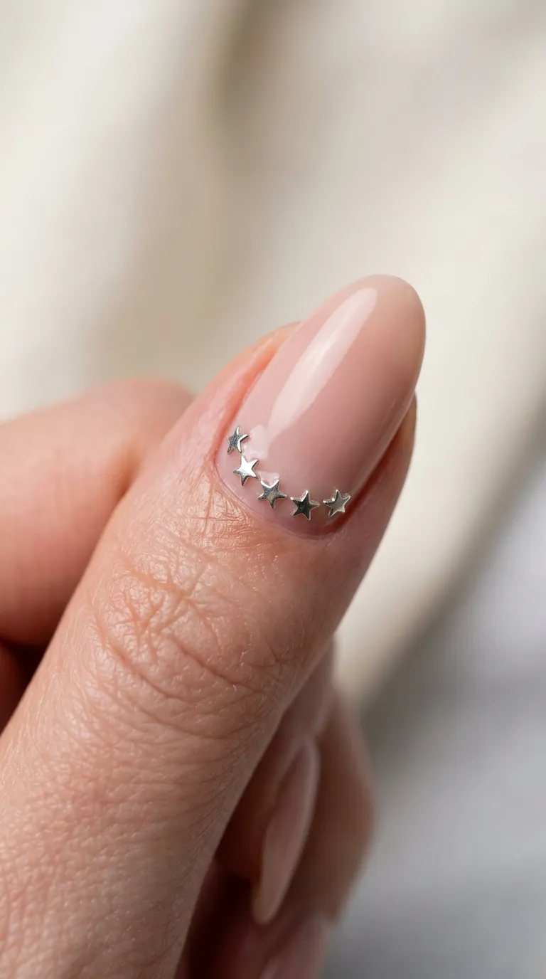 A delicate macro photograph of a single finger showing a nude pink nail with a trail of miniature silver star studs placed in a curved line following the cuticle shape. The focus is tight on the cuticle area showing precise placement of each star. Soft ring light creates even illumination and a tiny catchlight on each metallic star stud.