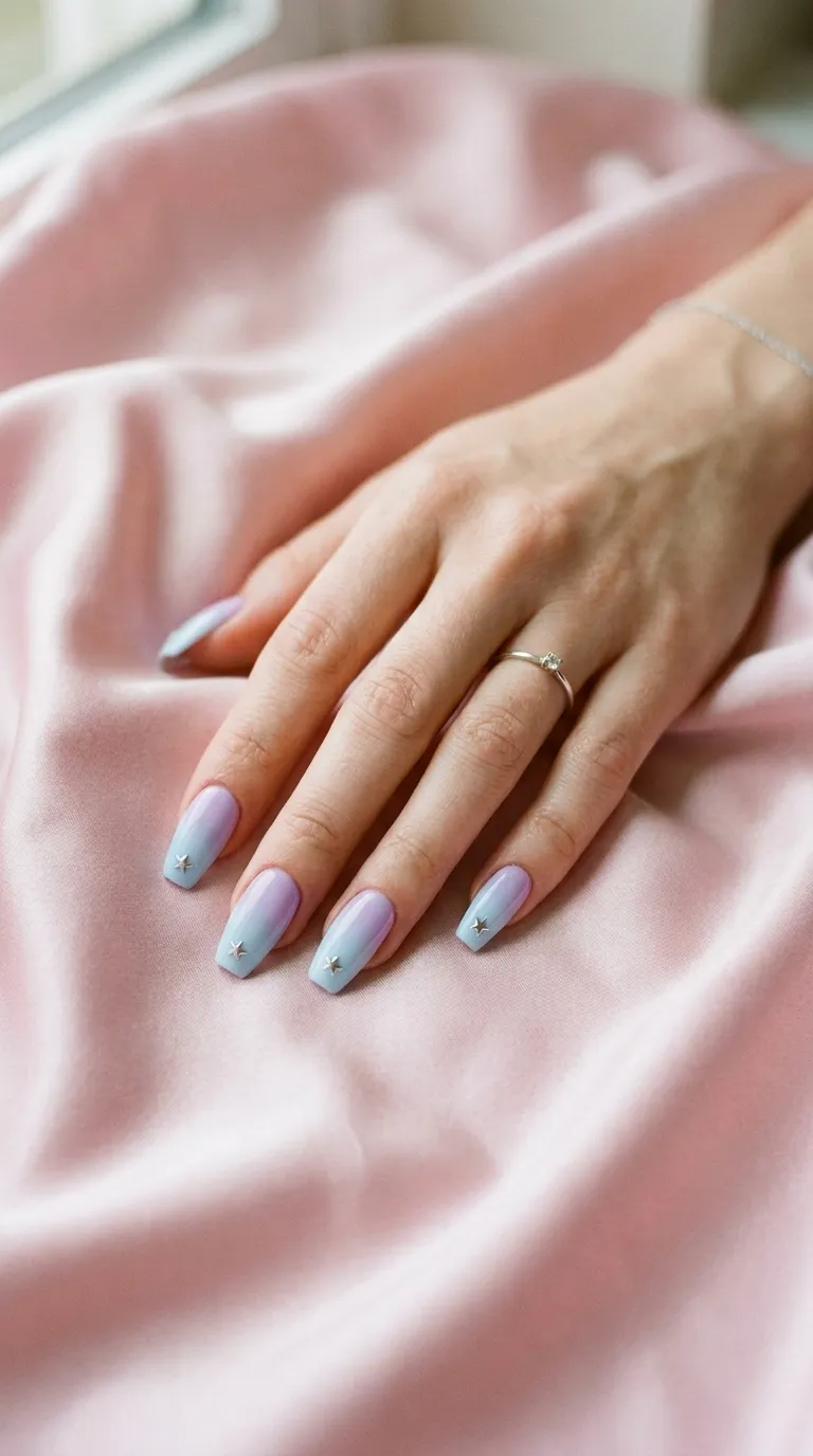 A softly lit photograph of a woman's hand displaying medium-length coffin-shaped nails with a smooth pastel gradient moving from lavender at the cuticle to baby blue at the tips. Miniature silver metallic star studs are placed near each nail tip. The hand rests on a pastel pink satin fabric with gentle wrinkles.