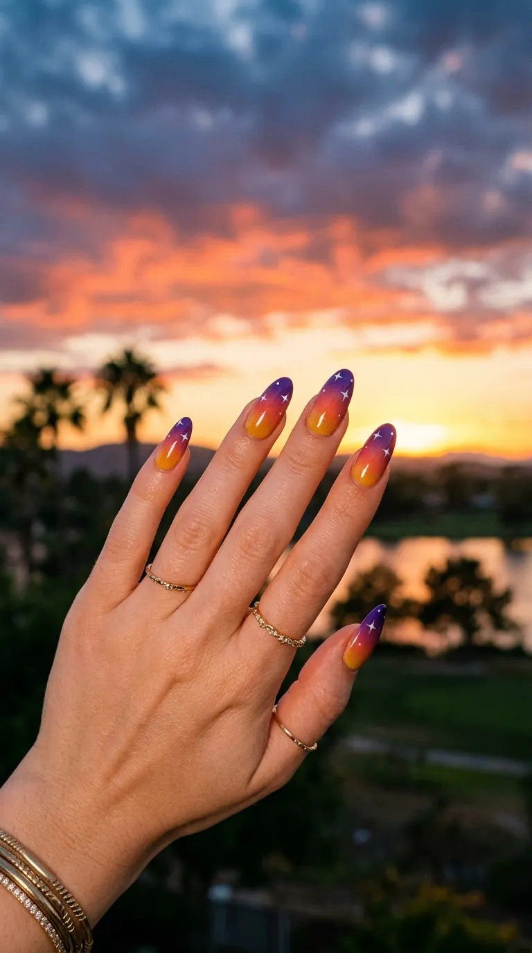 A warm photograph of long oval nails with a gradient ombre fading from golden yellow at the cuticle through orange and coral to deep purple at the tips. Small white star dots are scattered across the purple tip area. The hand is silhouetted against an actual sunset sky. Warm golden hour light, dreamy and atmospheric. Shot with a telephoto lens.