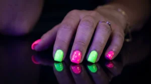 A close-up photograph of a woman's hand resting on a glossy black surface, her nails painted in vivid neon pink and electric green with tiny star shapes glowing under ultraviolet light. Each nail catches sharp reflections, and the background fades into deep shadow. Shot with a macro lens, shallow depth of field, studio lighting with a faint purple UV glow illuminating the neon pigments.